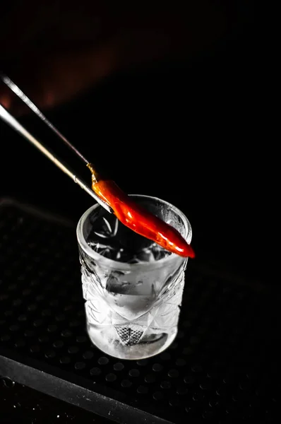 Bartender förbereder exotiska kryddig alkohol cocktail med chili paprika på bar på svart bakgrund — Stockfoto