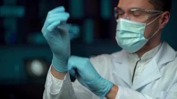 Close-up zicht op ernstige jonge bebaarde man arts in witte professionele jas het aantrekken van blauwe beschermende handschoenen over laboratorium achtergrond geïsoleerd — Stockvideo