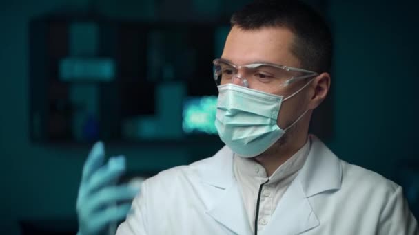 Beyaz profesyonel önlüklü, sakallı, ciddi genç bir doktorun laboratuvar geçmişine mavi koruyucu eldivenler giymesini yakından izleyin. — Stok video