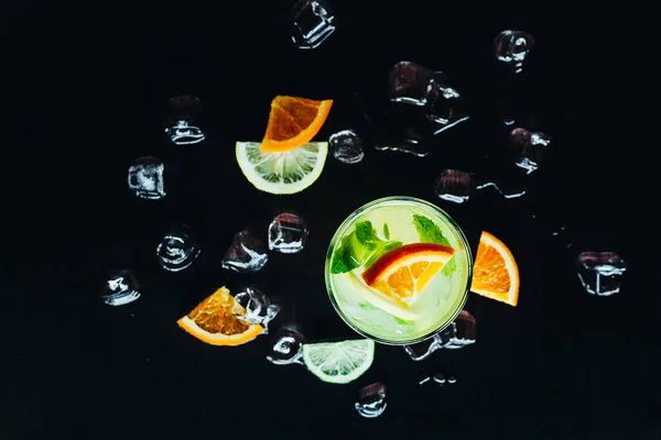 Top of view of glass with cocktail and ice with lime and orange slice on black background — Stock Photo, Image