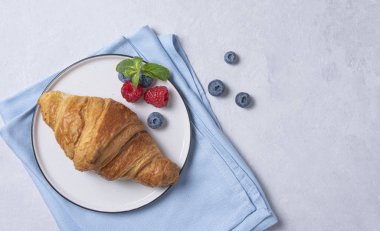 Beyaz tabakta taze kruvasan ve böğürtlenli mavi peçete.