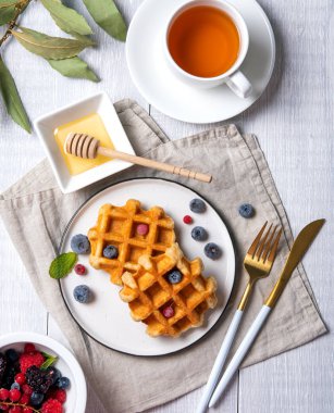 Böğürtlenli taze Belçika waffleları ve kırmızı frenk üzümü, bal ve çay beyaz bir masada. Nefis bir kahvaltı. Üst görünüm ve düz uzanma
