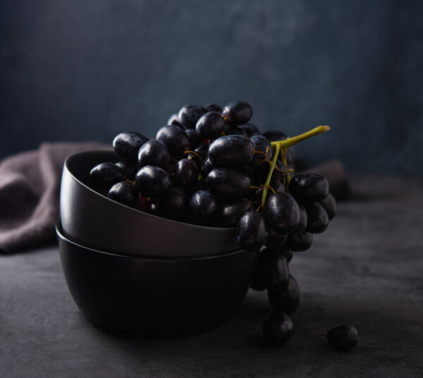 Группа сочного чёрного винограда лежит в тёмно-серой чаше на тёмно-сером столе. Фотография темная и настроение. Передний и макро вид
