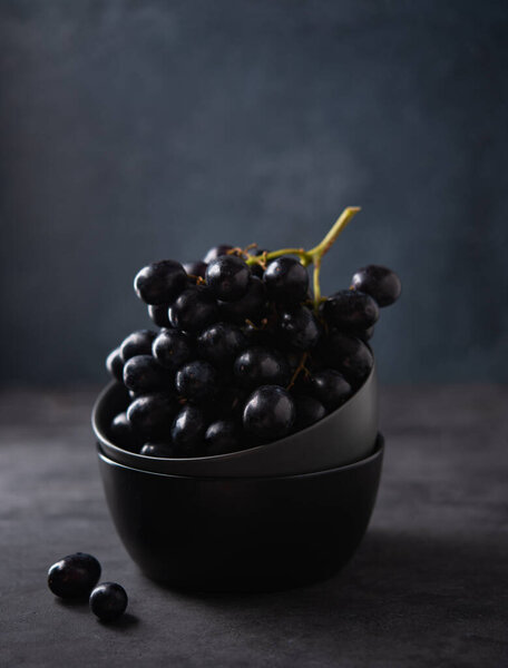 Группа сочного чёрного винограда лежит в тёмно-серой чаше на тёмно-сером столе. Фотография темная и настроение. Передний вид и пространство для копирования
