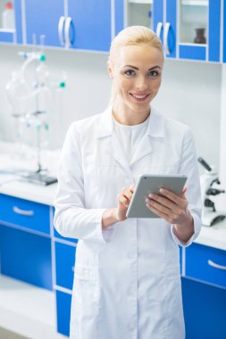 Yenilikçi tablet. Neşeli pozitif güzel kadın laboratuvarda ayakta ve modern tablet kullanırken labcoat giyiyor