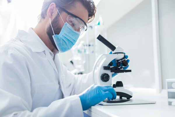 Modern lab. Smart nice handsome scientist sitting in front of the microscope and looking at the tablet screen while working on his research