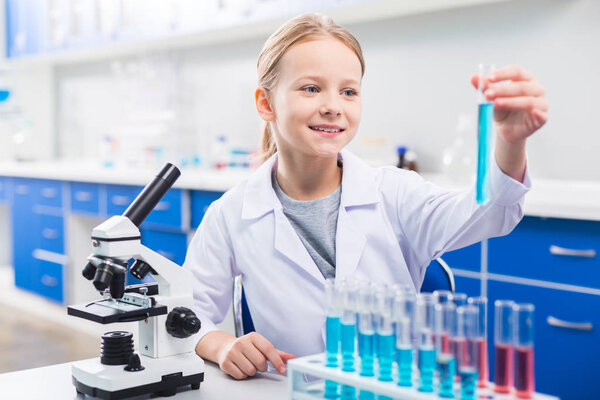 Blue liquid. Joyful sweet blond girl taking vial from stand while grinning and standing near microscope