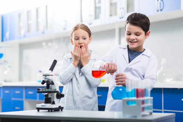 Dangerous experiment. Beautiful nice girl covering her mouth while looking at cheerful boy who combining liquids