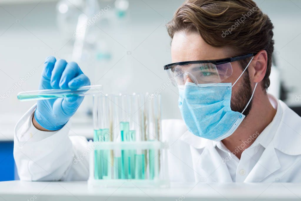 Professional chemist. Smart nice male researcher wearing a mask and protective glasses while conducting chemical experiment in the lab