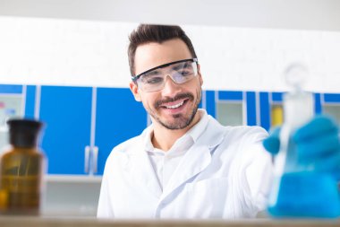 Artık gerek yok. Neşeli gay erkek laboratorian gülümseyerek ve ampul bakarken güvenlik gözlükleri giyiyor