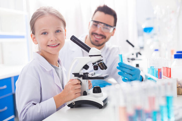 Exciting observation. Cheerful happy positive girl grinning while touching microscope and staring at camera