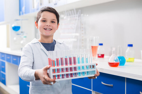 Little helper. Glad vigorous appealing boy holding stand with vials while gazing at camera and wearing lab coat