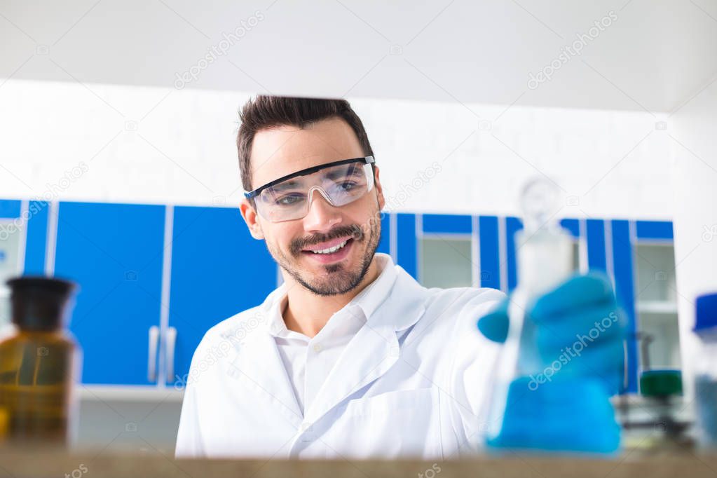 Creative method. Attractive bearded male laboratorian dressing in safety glasses while grinning and staring at bulb