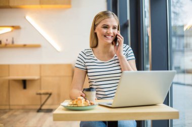 Mesafe iletişimi. Olumlu neşeli genç kadın kafede oturan ve dizüstü bilgisayar üzerinde çalışırken bir telefon görüşmesi yapıyor