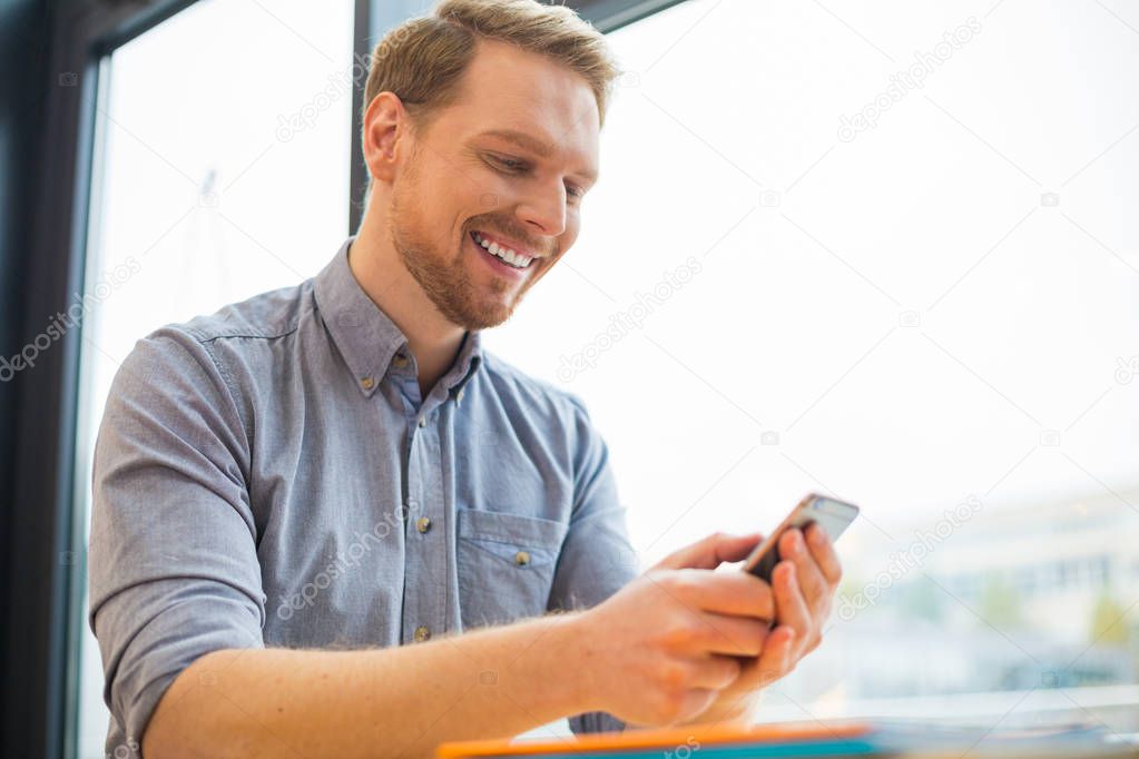 Modern technological device. Cheerful happy delighted man looking at the smartphone screen and smiling while using this device