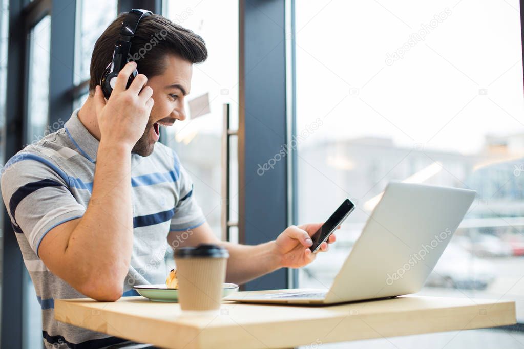 So exiting. Happy nice positive man holding his smartphone and expressing emotions while listening to music in the headphones