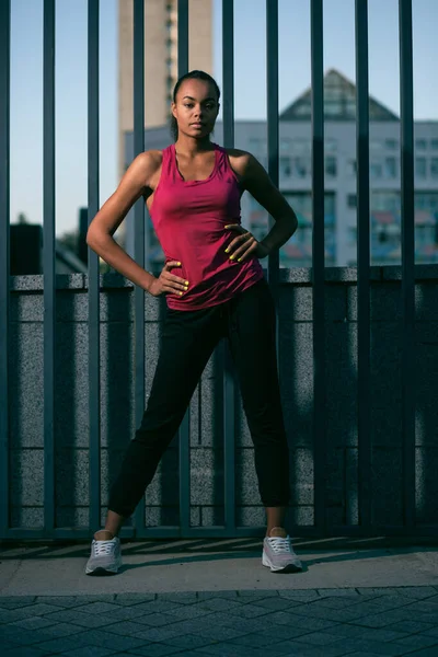 Calm confident sporty lady standing outdoors near the fence with her hands on the waist