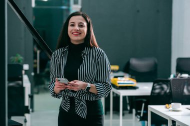 Ofisteki güzel genç bayan elinde modern bir akıllı telefon ve gülümsüyor.