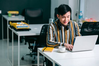 Genç gülümseyen adam ofiste dizüstü bilgisayarın önünde oturuyor ve mutlu görünüyor.