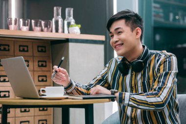 Masada dizüstü bilgisayarı ve bir fincan kahveyle oturan neşeli genç adam gülümseyerek yüzünü çeviriyor.