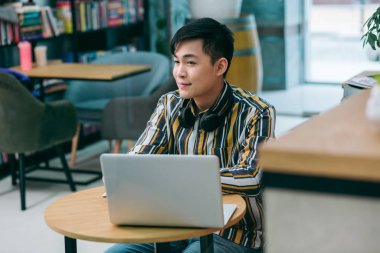 Modern bir dizüstü bilgisayarla masada oturan ve gülümseyerek yüzünü çeviren sakin genç adam.