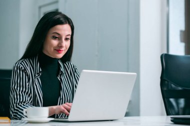 Gülümseyen ve modern bir dizüstü bilgisayarda çalışan kendine güvenen güzel genç bir kadın.