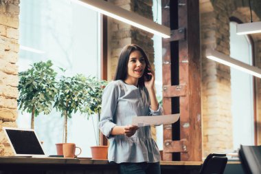 Elinde bir belgeyle durup telefonda konuşan neşeli bir kadın.