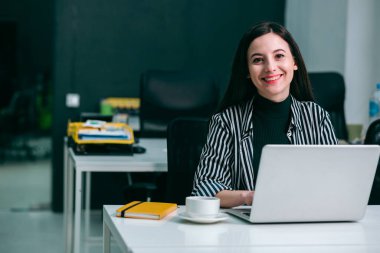 Ofiste dizüstü bilgisayarın önünde elinde bir defter ve bir fincan kahveyle oturan neşeli, güzel bir kadın.
