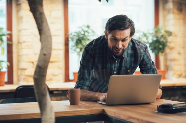 Gülümseyen genç adam dizüstü bilgisayarına eğiliyor ve ilgiyle ekrana bakıyor.