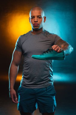 Genç profesyonel sporcu bir elinde koşu ayakkabısıyla duruyor ve ciddi görünüyor.