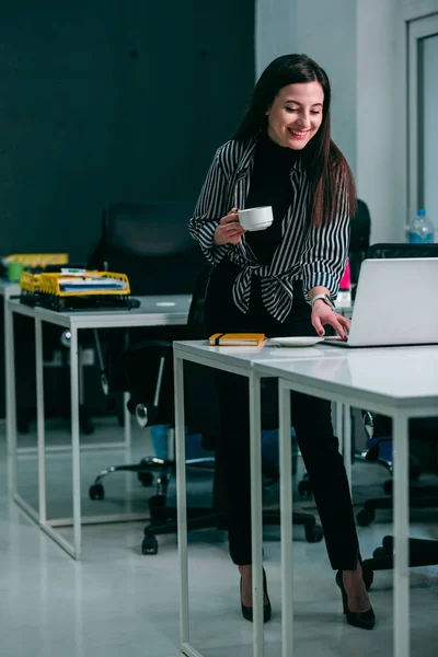 Mirthful lady standing with a cup and smiling while using a laptop