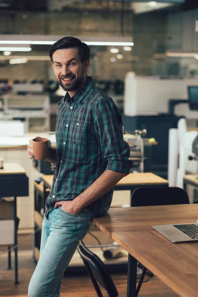 Handsome man in the office standing by the table with a mug and smiling