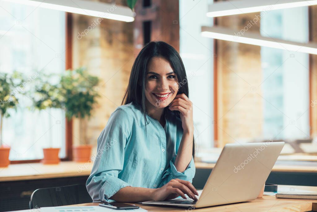 Cheerful young woman sitting at the table with a laptop and smiling while touching her face