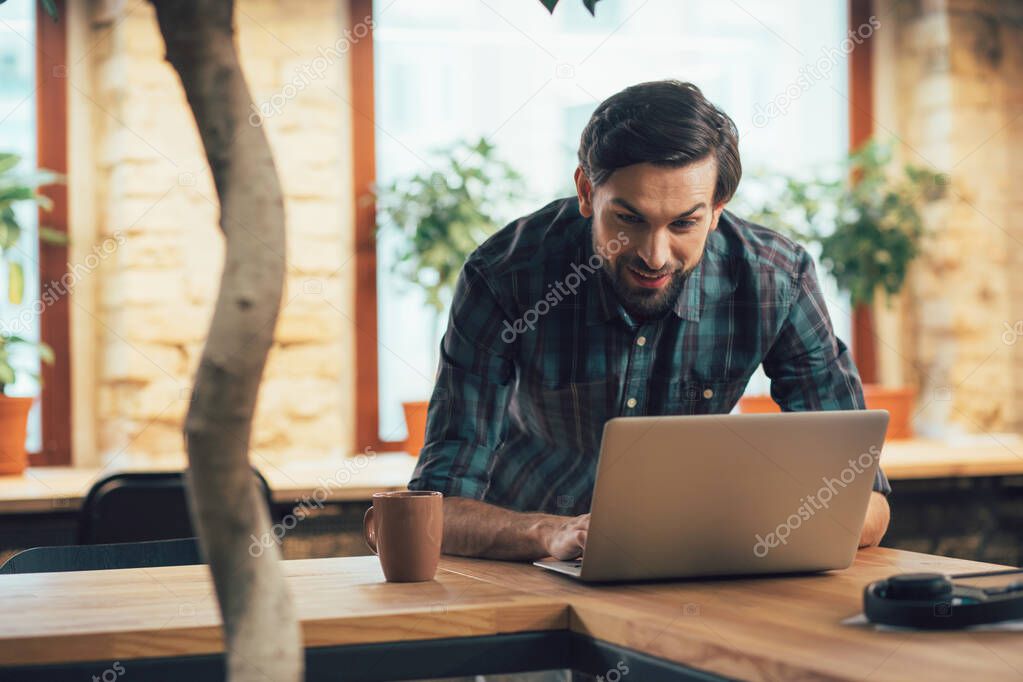Smiling young man leaning to his laptop and looking at the screen with interest