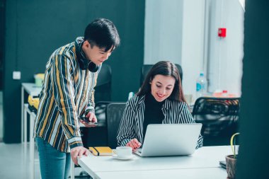 Gülümseyen bir kadın dizüstü bilgisayar kullanıyor ve yanında duran sakin bir iş arkadaşı ekrana bakıyor.