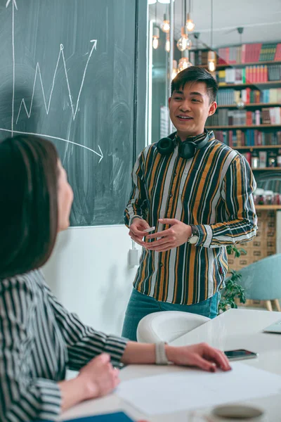 Cheerful young man standing in front of the company manager and showing a graphic on the blackboard