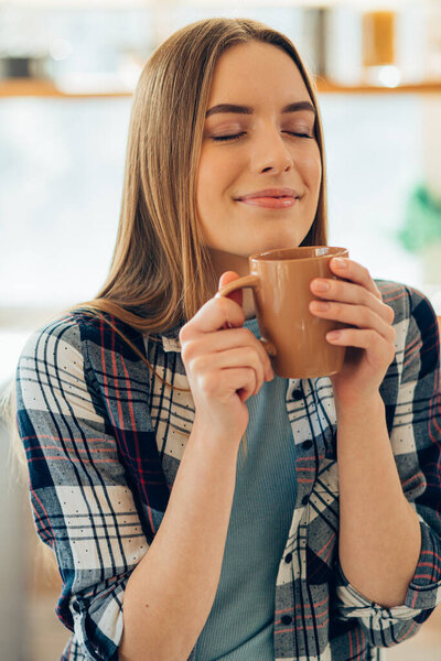 Pleased lady holding a mug near her face and closing her eyes while smelling the beverage