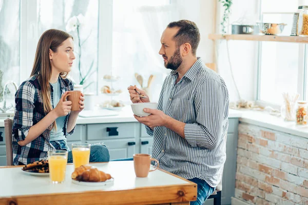 Young man eating cereals and frowning while looking at the young lady with tea in front of him