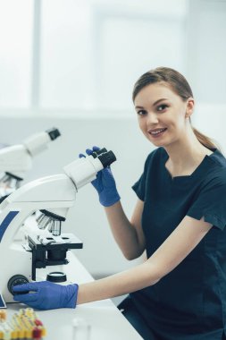Steril laboratuvarda çalışan pozitif bir bayan ve mikroskopla otururken gülümsüyor.