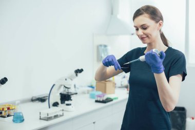 Lastik eldivenli neşeli laboratuar asistanı elinde mikroskop kaydırağıyla otomatik pipet makinesi kullanıyor.