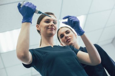 Neşeli laboratuvar çalışanı meraklı bir iş arkadaşının yanında duruyor ve otomatik pipet makinesi kullanıyor.