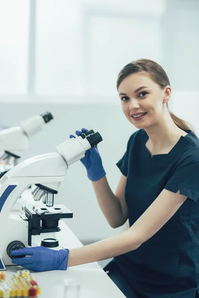 Positive lady working in sterile laboratory and smiling while sitting with a microscope