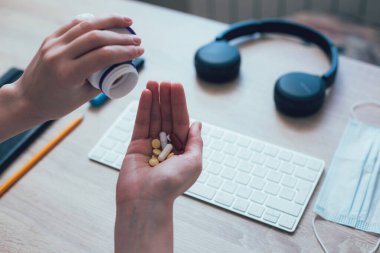 Masanın üzerinde renkli haplar olan bir elin kesilmiş fotoğrafı. Kablosuz klavye ve arka planda tıbbi maske.