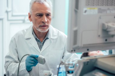 Ciddi sakallı bir doktor ultrason çevirici tutuyor ve ekran görüntülerine bakıyor.