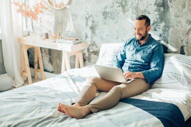 Yatakta dizüstü bilgisayarı olan neşeli genç bir adam. Ekrana bakarken gülümsüyor.