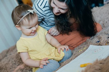 Neşeli kadın yeni yürümeye başlayan kızıyla oturuyor ve onunla çizim yaparken gülümsüyor.