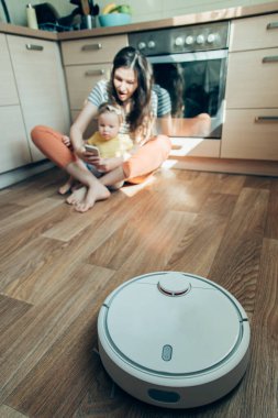 Modern robot süpürge mutfakta çalışıyor ve heyecanlı bir kadın kızıyla birlikte akıllı telefona bakıyor.