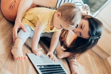 Yoğunlaşmış çocuk yerde oturup dizüstü bilgisayara dokunurken annesi arkasında oturuyor ve süreci kontrol ediyor.