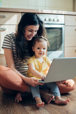 Mutfakta dizüstü bilgisayarla oturmuş gülümseyen bir kadın ve mutlu kızı bacaklarının üzerinde oturuyor.