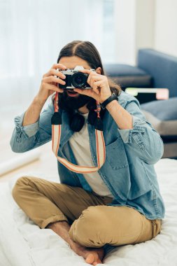 Gündelik giysiler içinde genç bir adam evde bir yatakta oturup fotoğraf çekiyor.